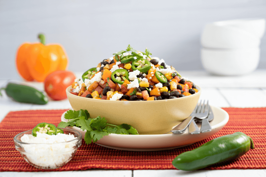 The image shows a red woven placemat with a white plate and a white bowl on top of the plate. There are three forks on the plate beside the bowl. In the bowl is a colourful medley of chopped tomatoes, corn, black beans, fresh jalapeno peppers, and feta cheese. The upper right of the image in the background shows three stacked white bowls out of focus. The lower right of the image shows a whole fresh green jalapeno pepper. The lower left of the image shows a small clear glass bowl filled with feta cheese and two sliced of fresh green jalapeno. In the upper left of the image, out of focus in the background is an orange pepper, tomato, and green jalapeno pepper.