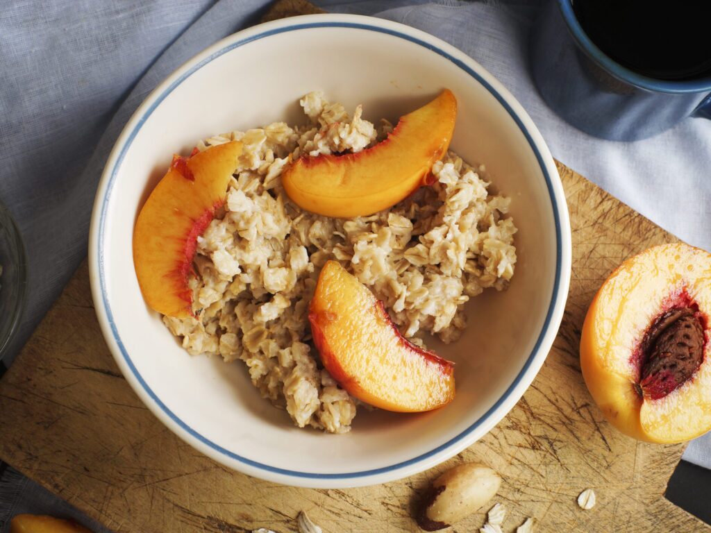 A bowl of oatmeal with sliced peaches on a wooden table. The oatmeal is light brown and topped with milk. The peaches are orange and sliced.