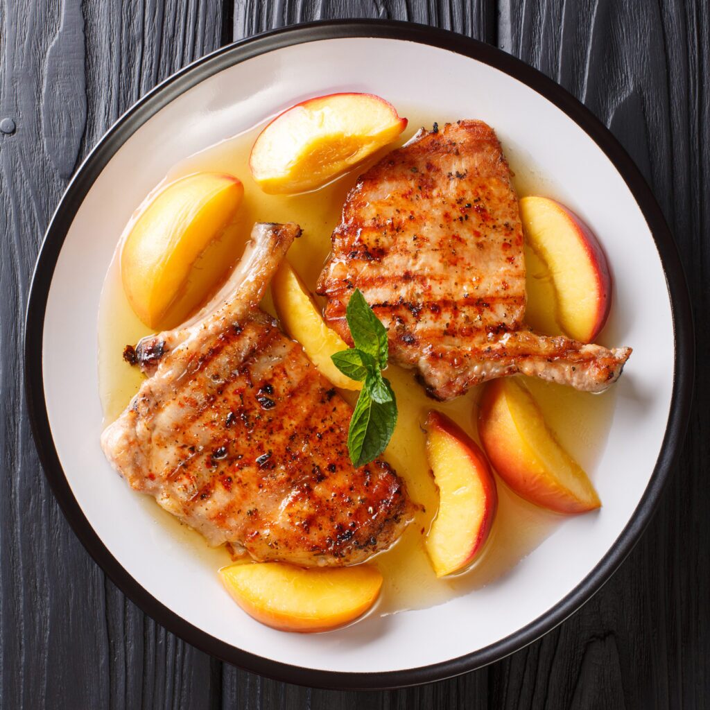 A plate of grilled pork chops and glazed peaches. The two pork chops are bone-in and have a browned crust. They are placed side-by-side on a white plate. To the left of the pork chops are two halved peaches. The peaches are a yellow-orange colour and appear ripe with a slight reddish hue. They appear to be glazed and and have grill marks. A sprig of fresh mint rests on top of the pork chops. In the background, slightly blurred, is a brown wooden table.