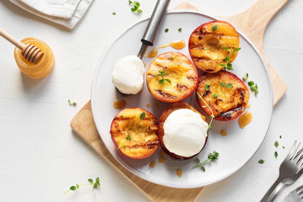 A plate of three grilled peach halves topped with scoops of vanilla ice cream. The peaches are golden brown with grill marks and the ice cream is melting slightly. A drizzle of honey is on each peach half and there are sprigs of fresh thyme scattered around the plate.