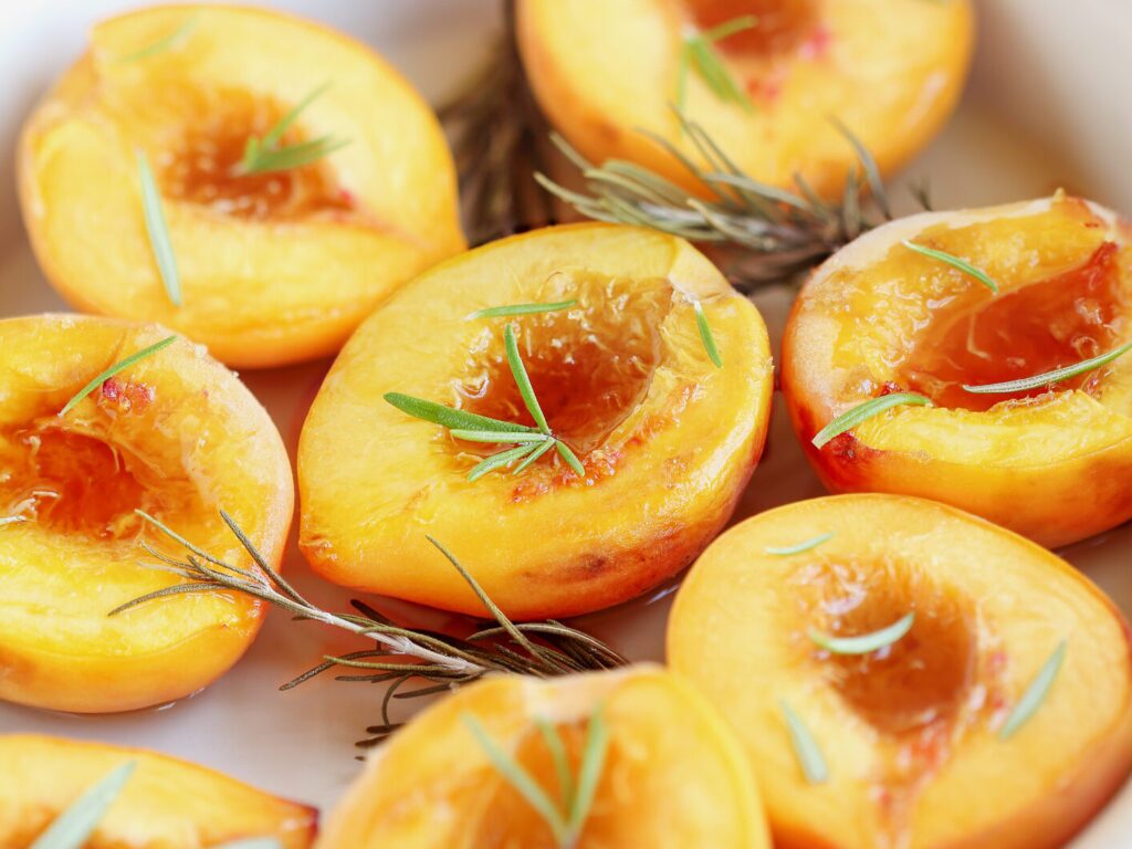 A plate of roasted peaches with thyme and brown sugar. The peaches are halved and golden brown.  There are sprigs of fresh thyme on the plate and brown sugar is sprinkled around the peaches.
