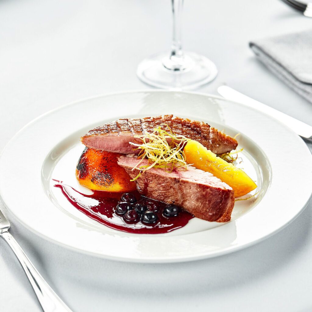 A white plate with a grilled duck breast sliced in half.  Next to the duck is a half of a grilled peach.  Both the duck and peach are drizzled with a dark red berry sauce.  There are also a few green leaves for garnish.