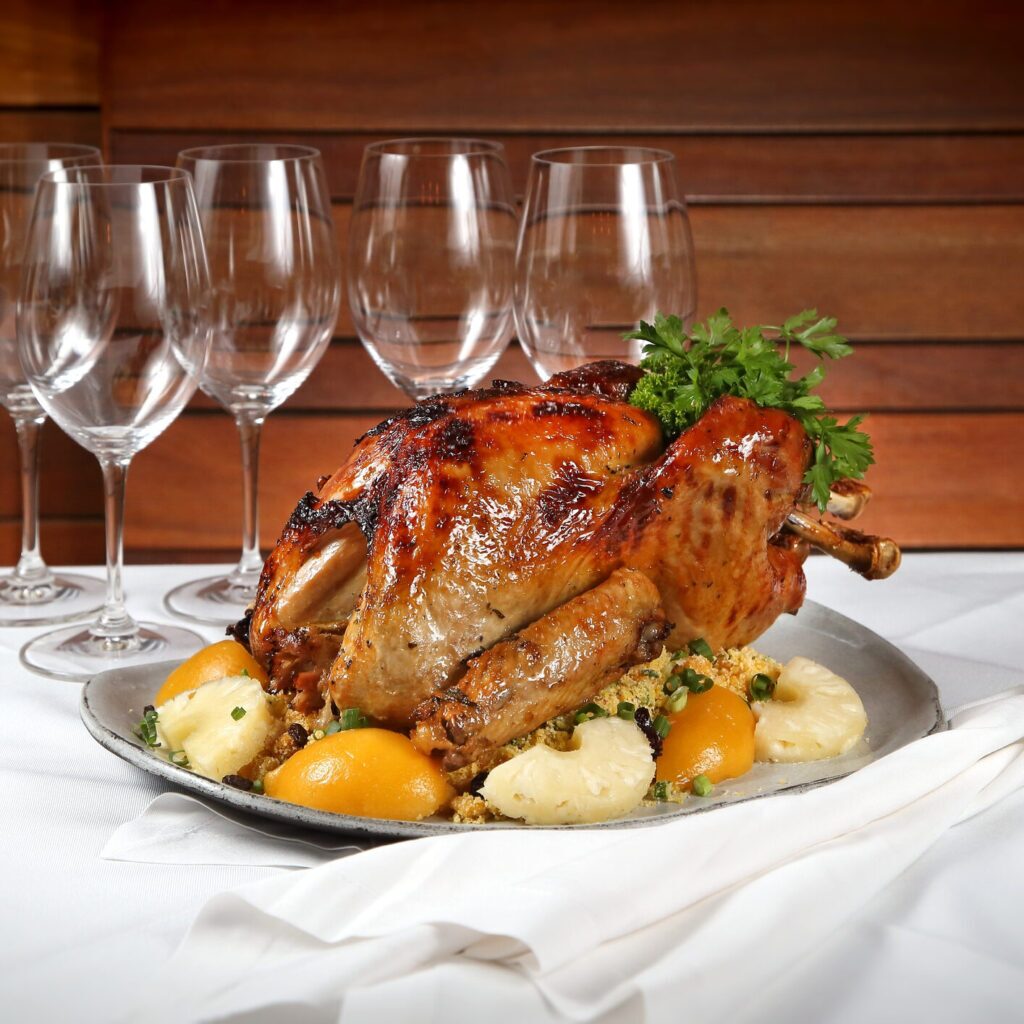 A roasted whole turkey on a platter. The turkey is brown and crispy, and there is fruit garnish around the base. In the background, there are wine glasses on a table.