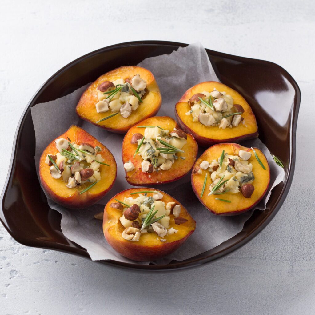A plate of blue cheese stuffed peaches with nuts and rosemary. The peaches are halved and roasted a golden brown. The blue cheese filling is visible in the center of each peach half. There are chopped nuts and rosemary sprigs on top.