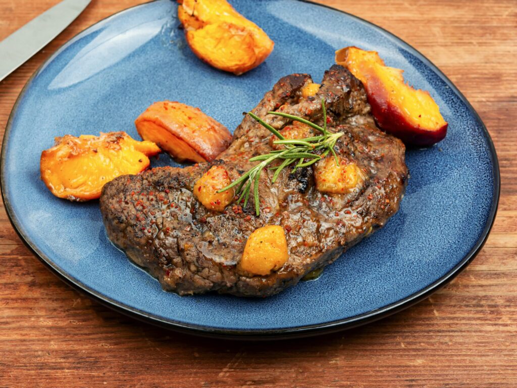 A blue plate with a beef steak topped with roasted peaches. The steak is sliced and cooked medium-rare. The peaches are golden brown with grill marks. There is a brown sauce drizzled over the steak and peaches.