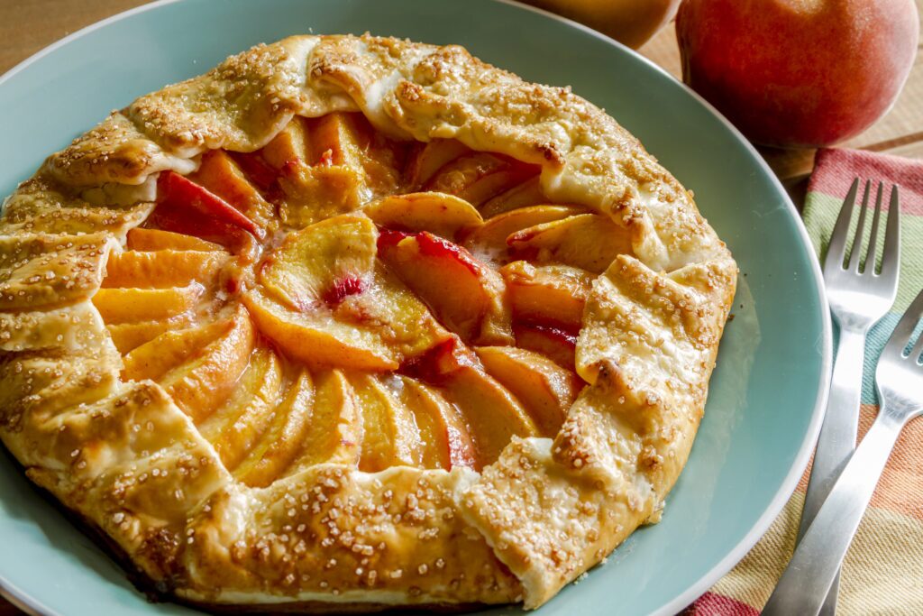 An overhead view of a French peach galette on a teal plate. The galette features a rustic, golden-brown crust with sugared edges, encasing a beautifully arranged filling of sliced peaches, showing a gradient of yellow to red hues.