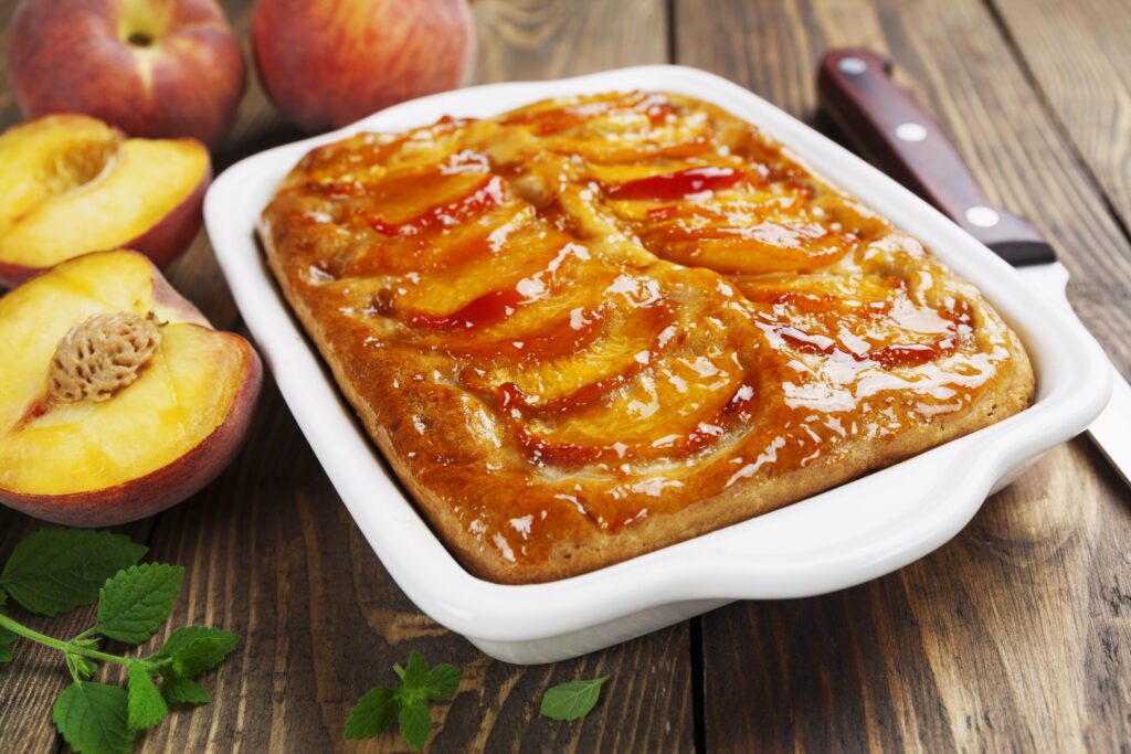 A square casserole dish made of clear glass filled with a peach pie filling. The pie filling is visible through the glass and appears to be a thick, chunky mixture of sliced peaches with a golden brown crust on top. The casserole dish is sitting on a wooden table with a light brown stain.  In the top left corner of the image, there is a  label that reads  “Casserole Dish Peach Pie”.