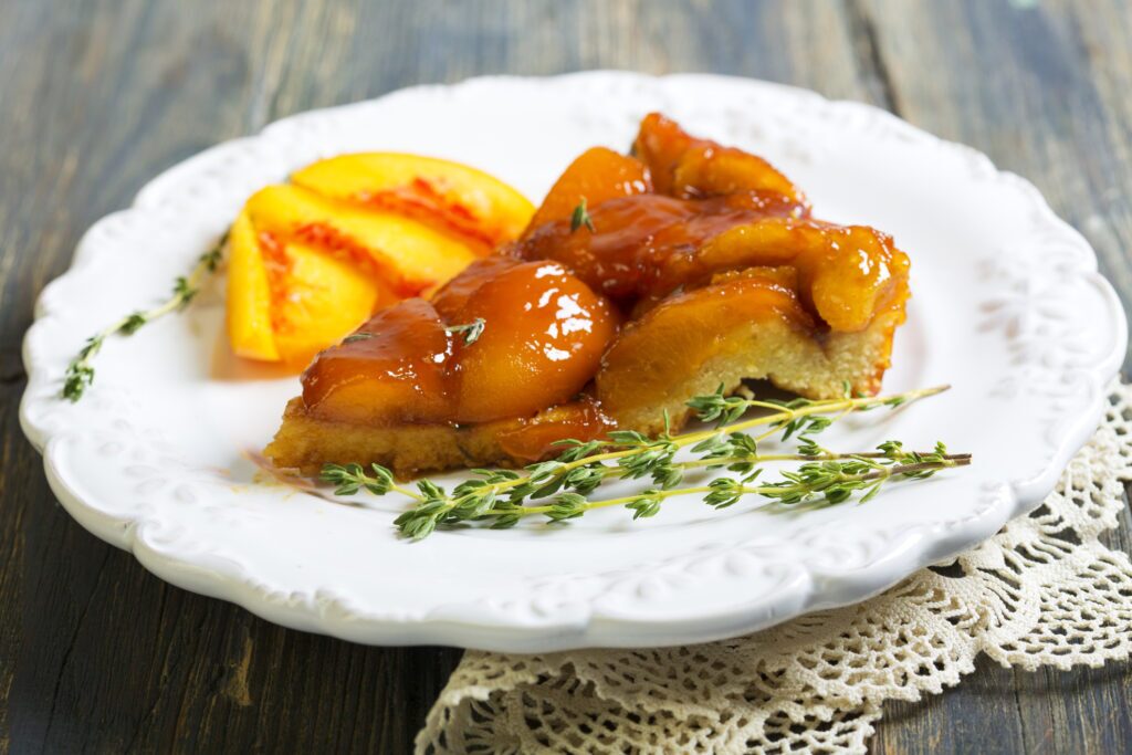 A slice of peach tart on a white plate. The tart crust is visible around the edge of the slice and appears to be golden brown and flaky. The peach slices on top of the tart are arranged in a fan shape and are light orange in color. There are a few sprigs of fresh thyme scattered around the peach slices. I