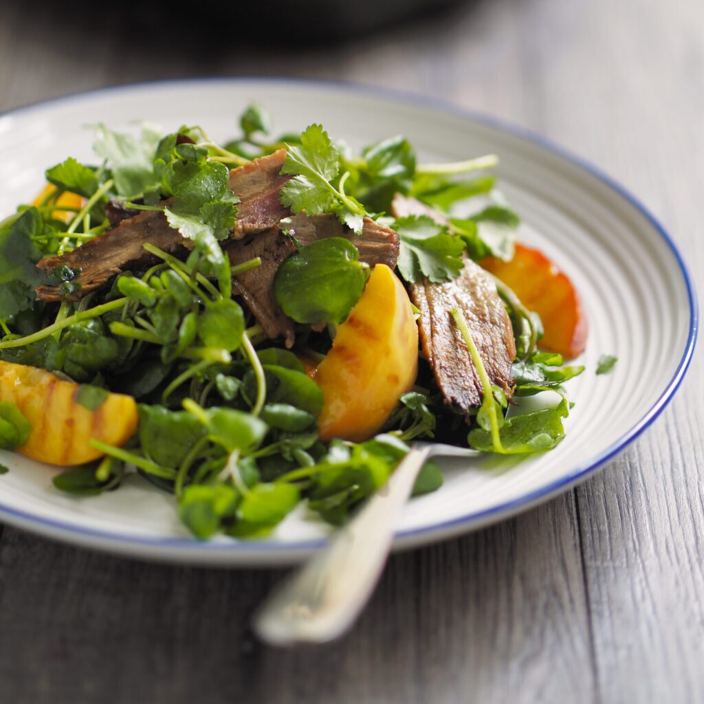 A white plate topped with a salad. The salad consists of a bed of watercress, slices of grilled duck breast, and grilled peach slices. A silver fork rests on the side of the plate. 