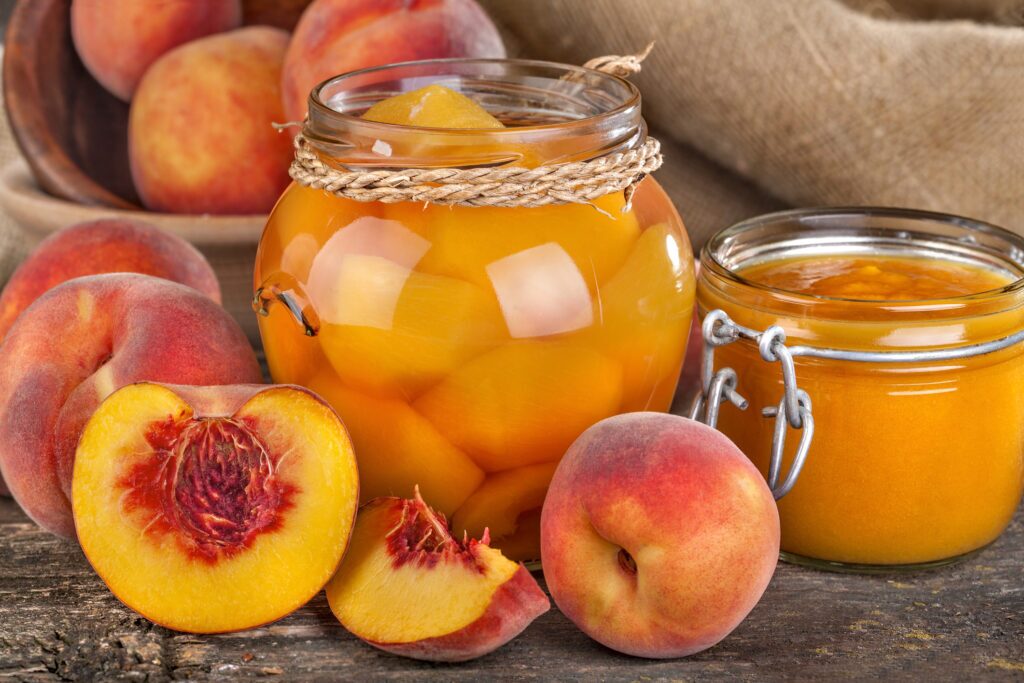 A glass jar filled with yellow peaches submerged in a clear liquid. The jar is sealed with a metal lid and has a label. The label appears to be brown and gold with text written on it. The table the jar is sitting on is wooden with a brown stain.