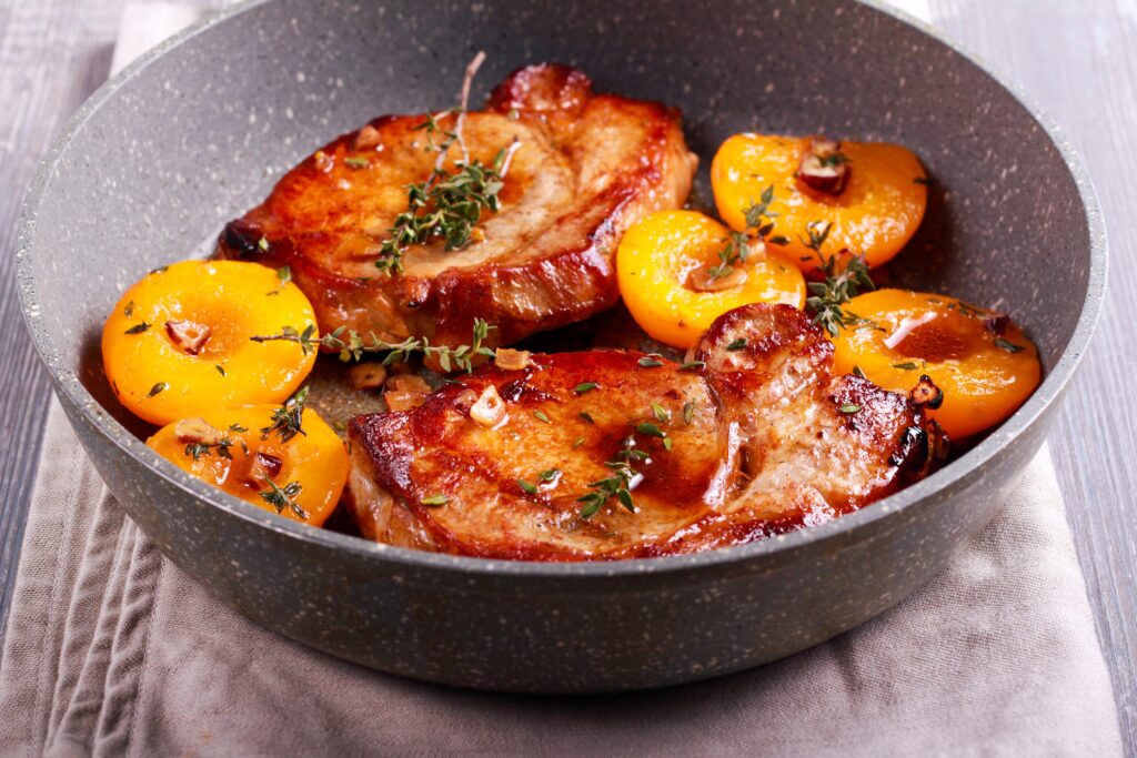 A frying pan containing two pork chops with sprigs of thyme and halved peaches. The pork chops are a light brown colour and appear to be cooked. The peaches are a yellow-orange colour and some appear ripe with a slight reddish hue. They are cut in half and appear to be seared on the cut side. There is a small amount of cooking liquid in the pan, and the thyme sprigs are scattered around the pan. 