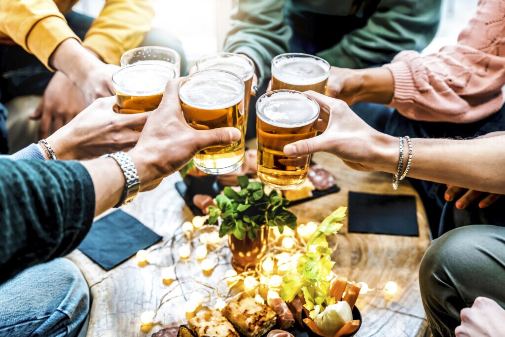 Friends cheering beer glasses on wooden table at brewery bar indoor at patio party - People having fun at happy hour at bar restaurant - Food and beverage lifestyle concept