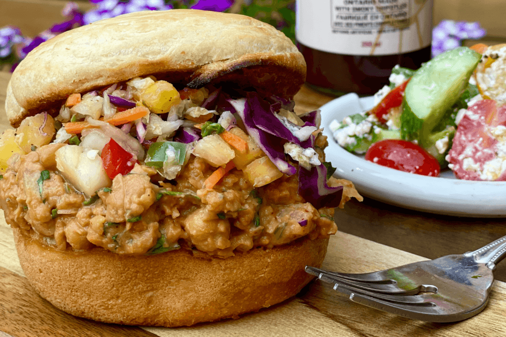 The image shows a closeup of a toasted burger bun with a chickpea mixture and colourful slaw. The sandwich is on wooden table. To the right of the sandwich is a white plate with tomatoes, red peppers, and sliced cucumber mixed with feta cheese. The top of a fork is visible in the foreground of the image.