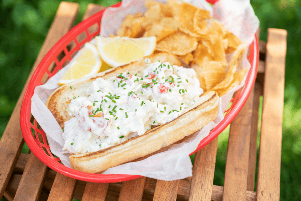 Lobster Roll. The image shows a slatted wooden table with a red plastic fry basket on it. In the fry basket is a hot dog bun loaded with lobster meat, mayonnaise, and a sprinkle of fresh chives. Atop the roll are two wedges of lemon and beside it are Farm Boy Crinkle Cut Potato Chips