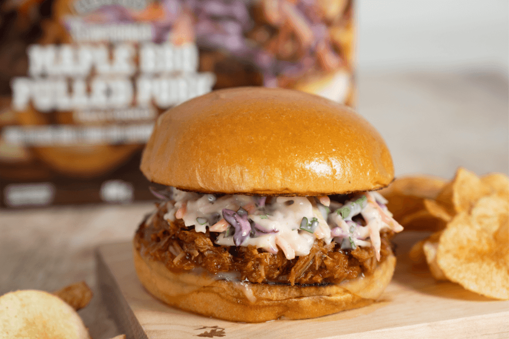 The image shows a pulled pork sandwich on a wooden cutting board next to potato chips. The sandwich is on a golden brioche bun and is topped with creamy coleslaw.