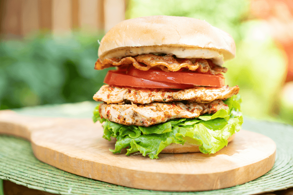 The image shows a grilled chicken BLT sandwich on a wooden cutting board. The sandwich is stacked with lettuce, grilled chicken, tomato slices, and crispy bacon on a burger bun.