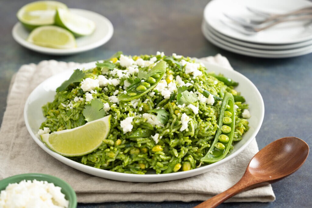 The image shows a white dish on a linen cloth. The dish has a green vegetable rice dish and is garnished with crumbled feta cheese and a wedge of lime.