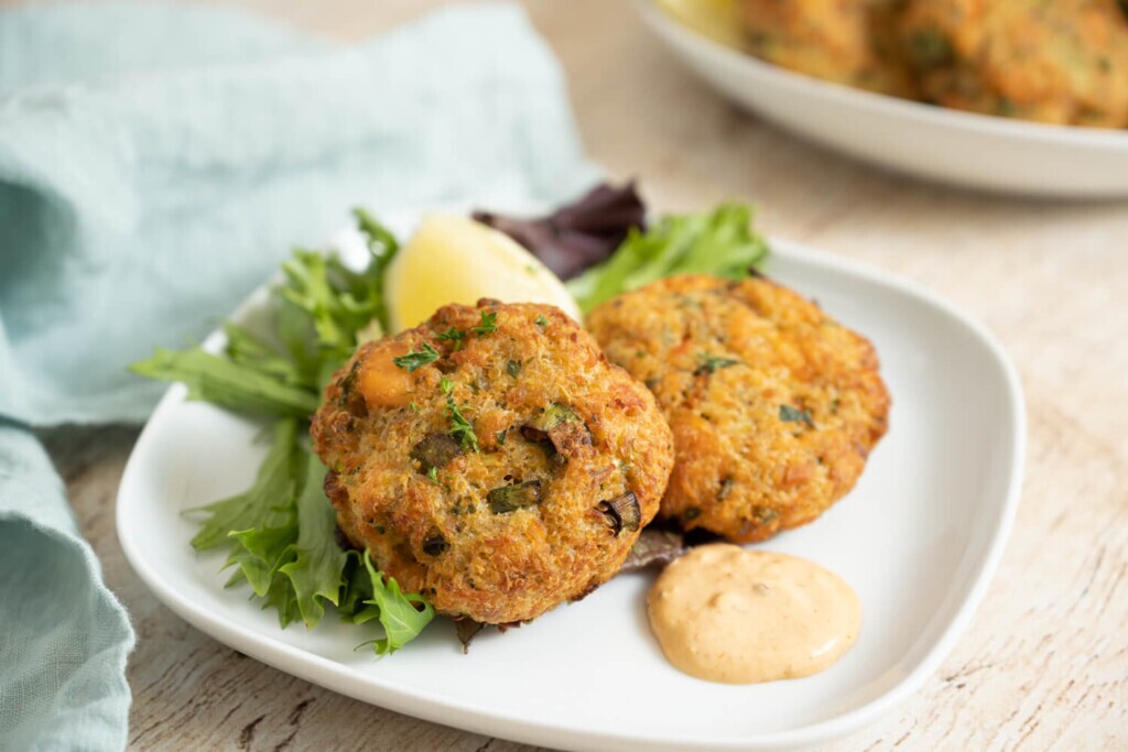 Leftovers Recipe: The image shows a rounded square plate with two salmon quinoa cakes made using leftover salmon. They are on a bed of mixed lettuce and garnished with a wedge of lemon and Farm Boy Chipotle Mayonnaise.