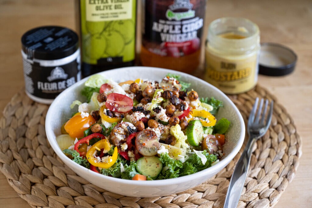 Angled view of a white bowl filled with salad in front of Farm Boy Fleur de Sel, Farm Boy Extra Virgin Olive Oil, Farm Boy Organic Apple Cider Vinegar, and Farm Boy Dijon Mustard