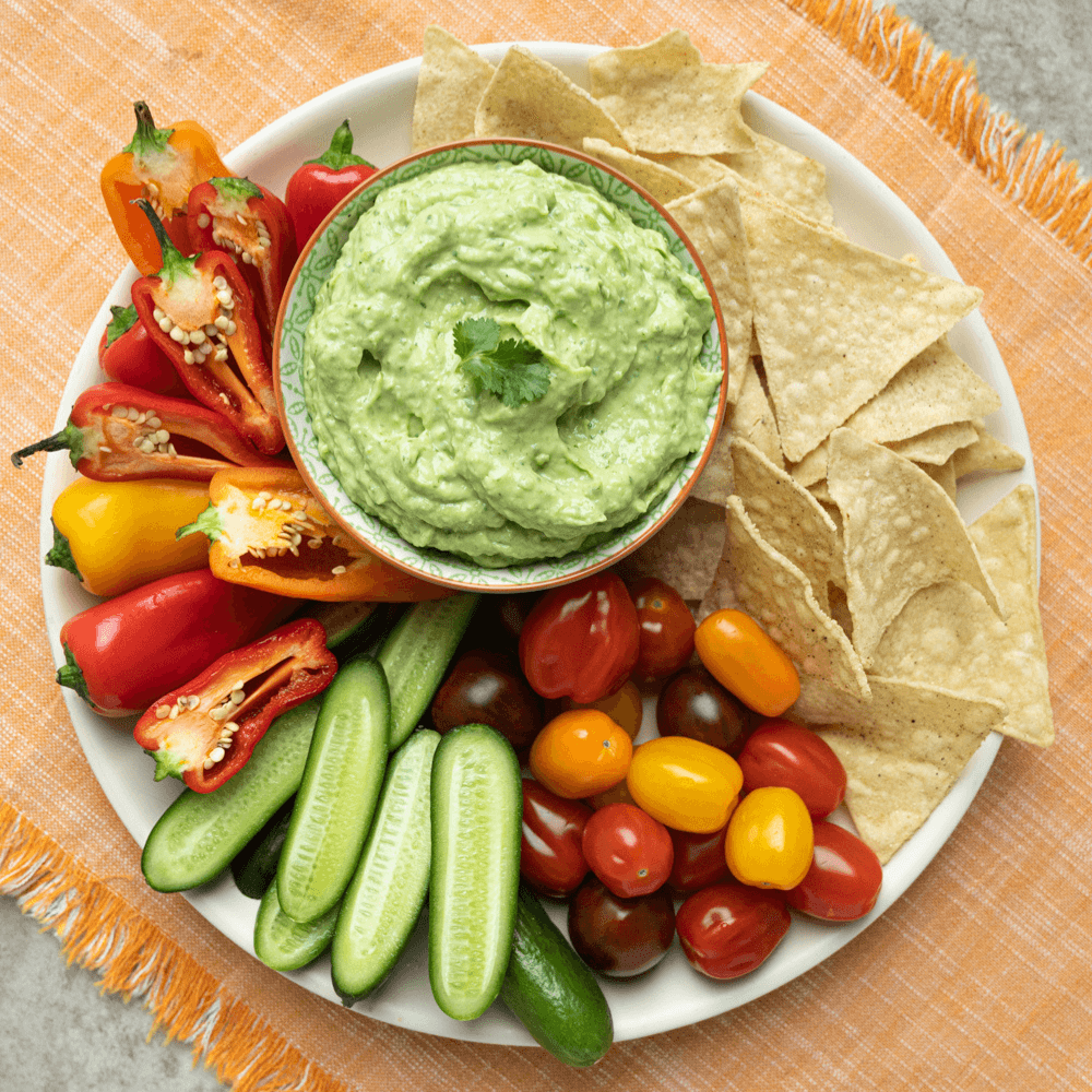 Overhead image of a white plate filled with fresh vegetables and tortilla chips. In the centre is a bowl of creamy avocado dip.