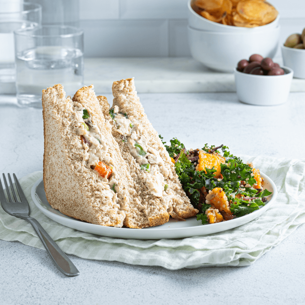 White plate with two pieces chicken salad sandwich on it, standing upright, with a kale salad beside it.