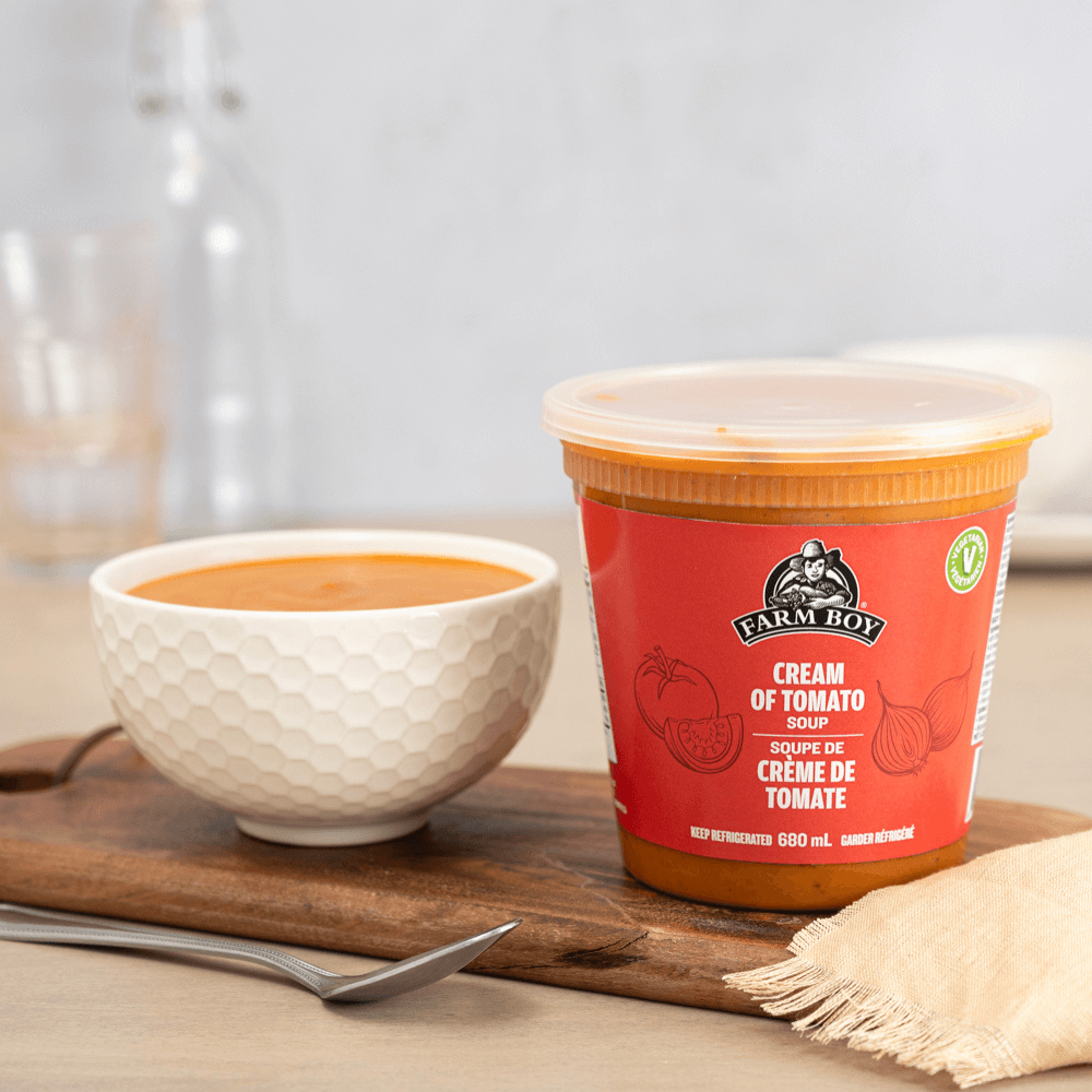 Farm Boy™ Creamy Tomato Soup container and white bowl filled with soup on dark wood cutting board with spoon in front.