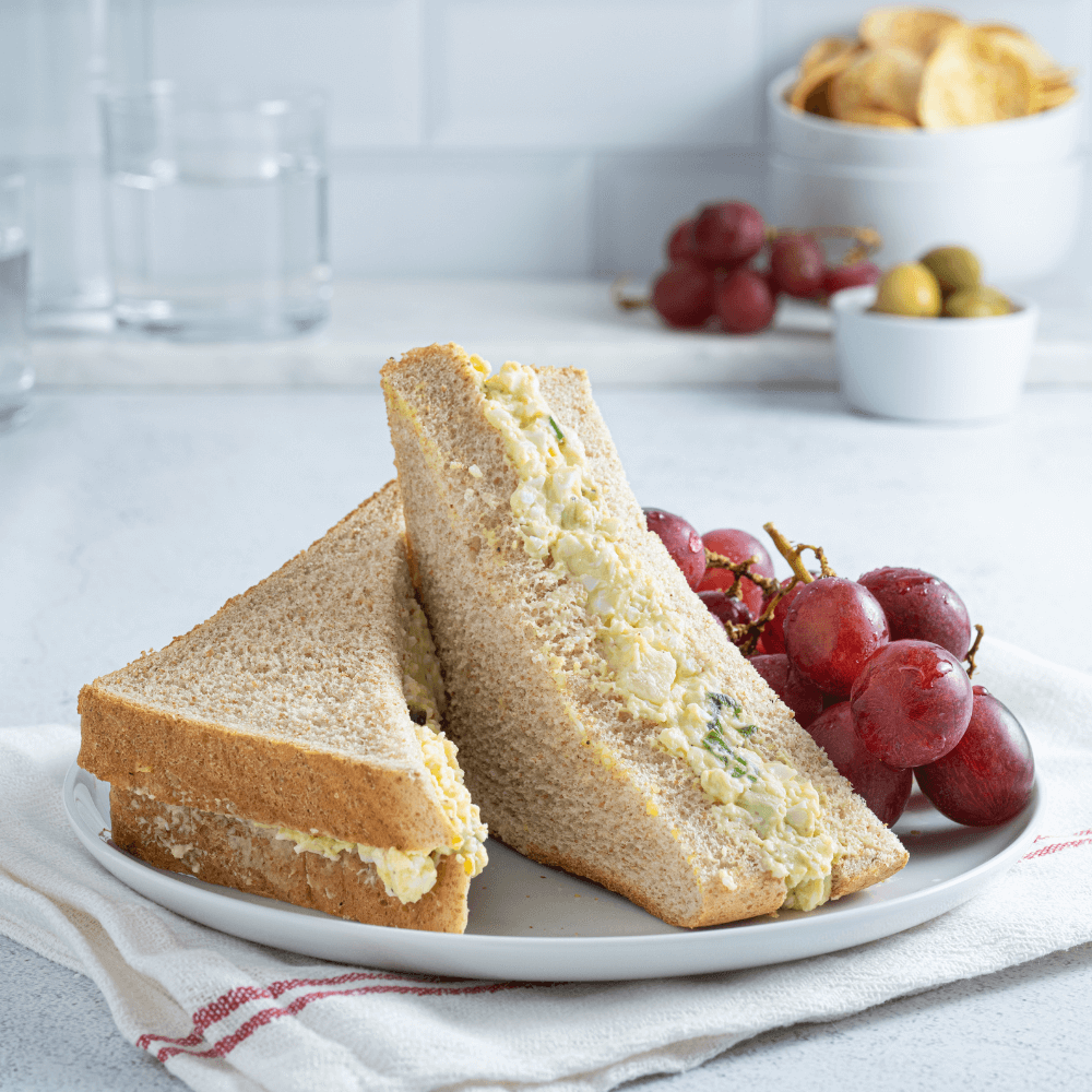 White plate with two halves of an egg salad sandwich on wheat bread with a small bunch of grapes beside it.