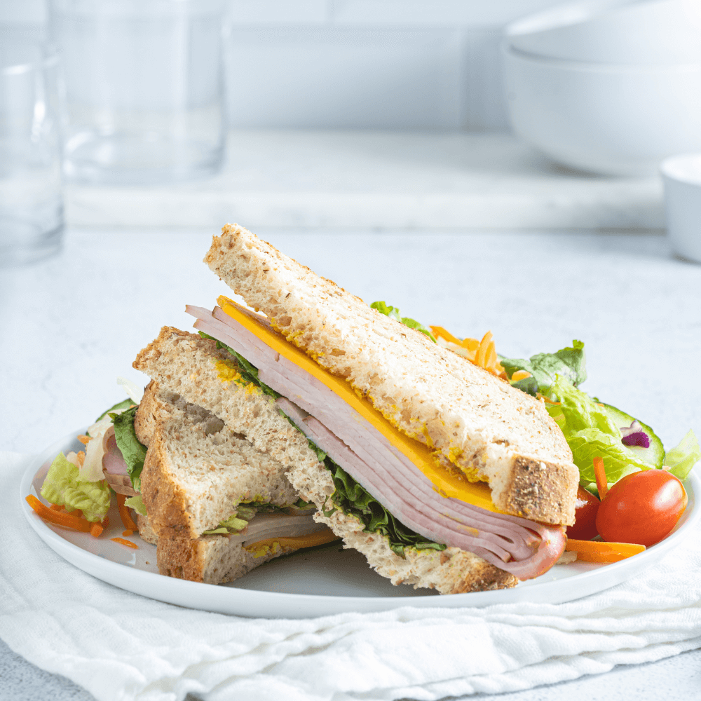 White plate with two halves of a ham and cheddar cheese sandwich. Behind it is a fresh garden salad.