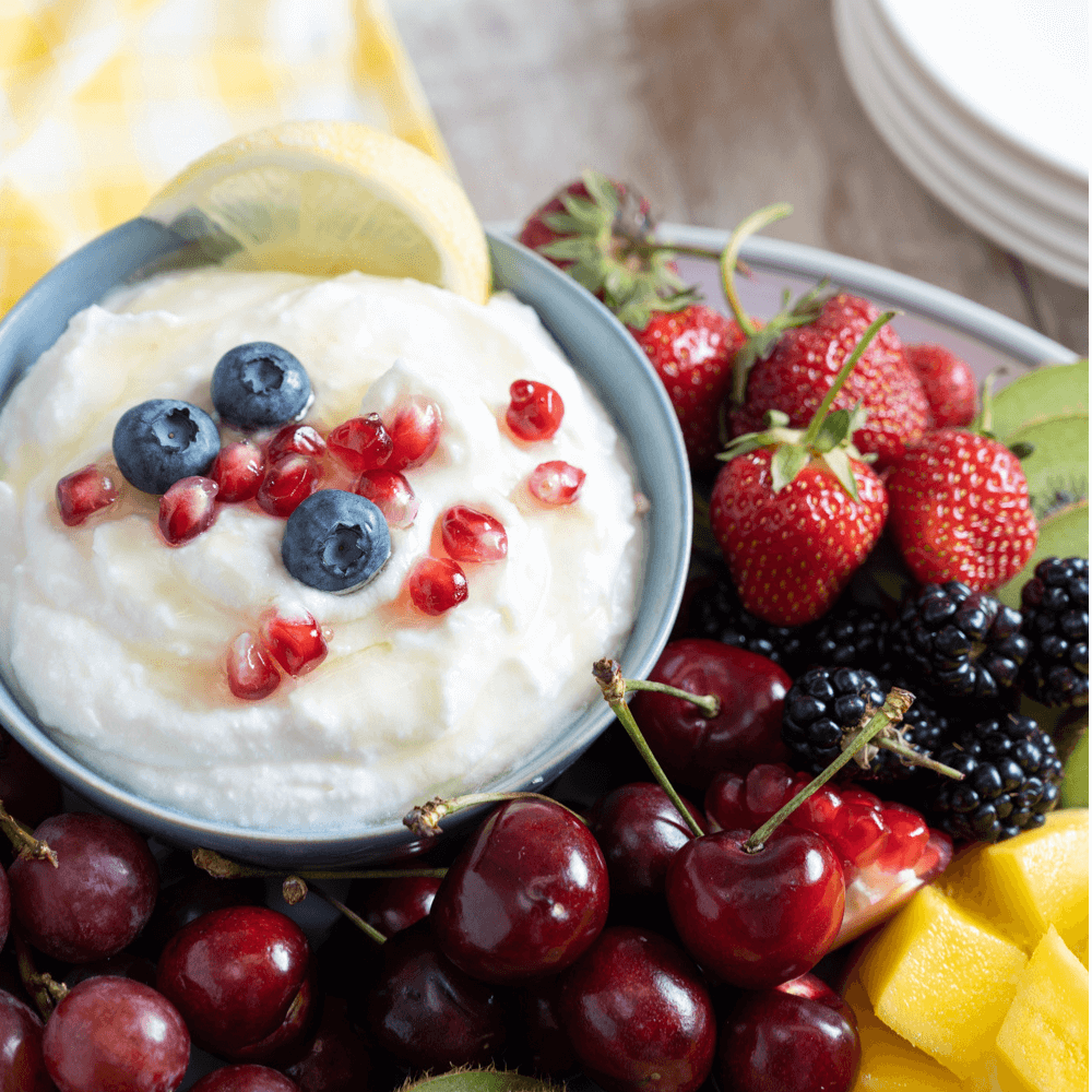 Small blue bowl filled with whipped honey lemon ricotta garnished with pomegranate arils and blueberries on a plate of assorted fresh fruits.