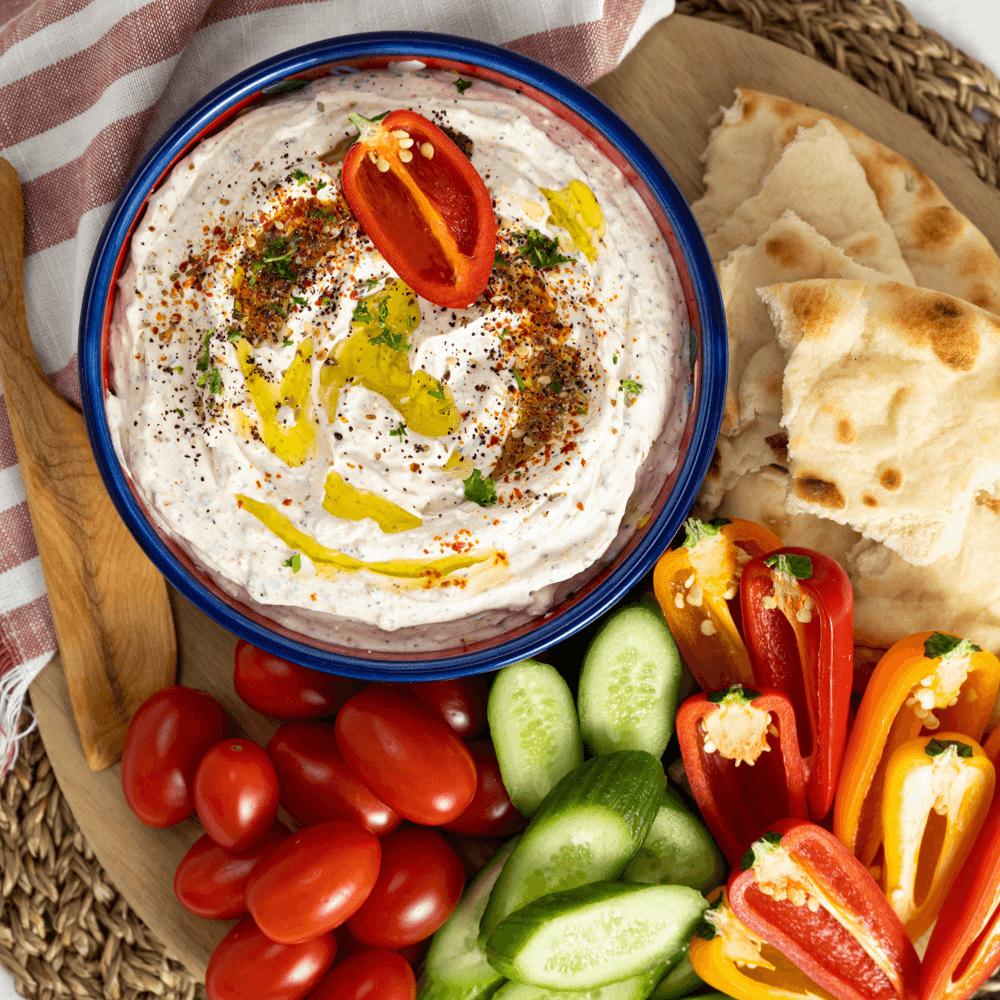 Overhead image of a blue-rimmed bowl filled with a spiced ;abneh dip. Next to the bowl are cherry tomates, sliced peppers, sliced cucumbers, and torn naan bread.