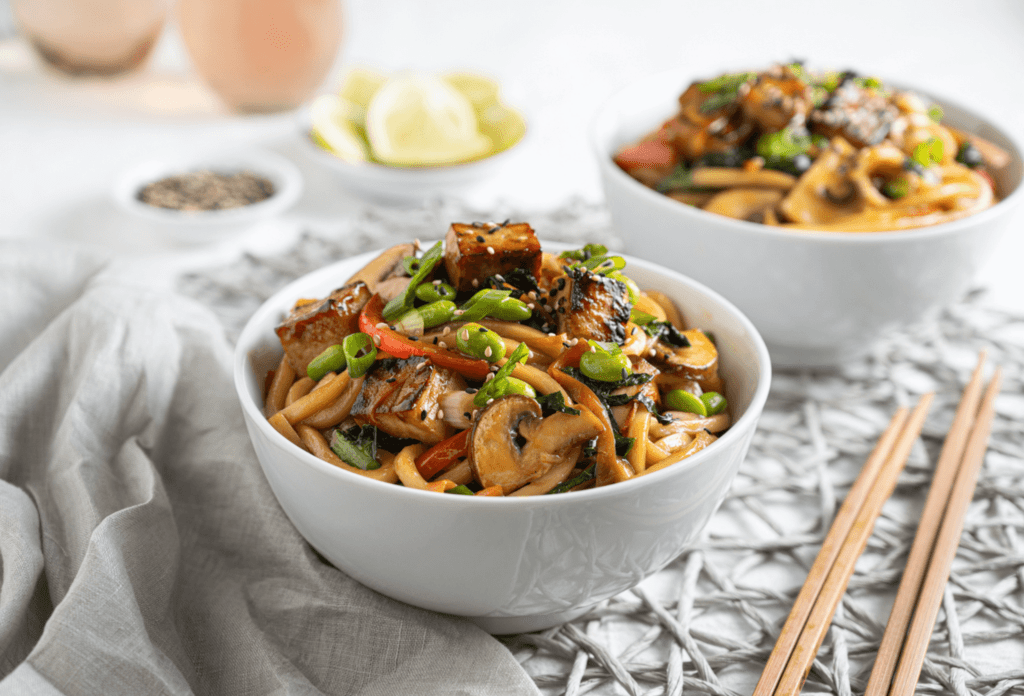 Vegan Recipe: A white bowl filled with teriyaki tofu and noodle stir fry. Another identical filled bowl is in the background. A pair of chopsticks is beside the bowl.