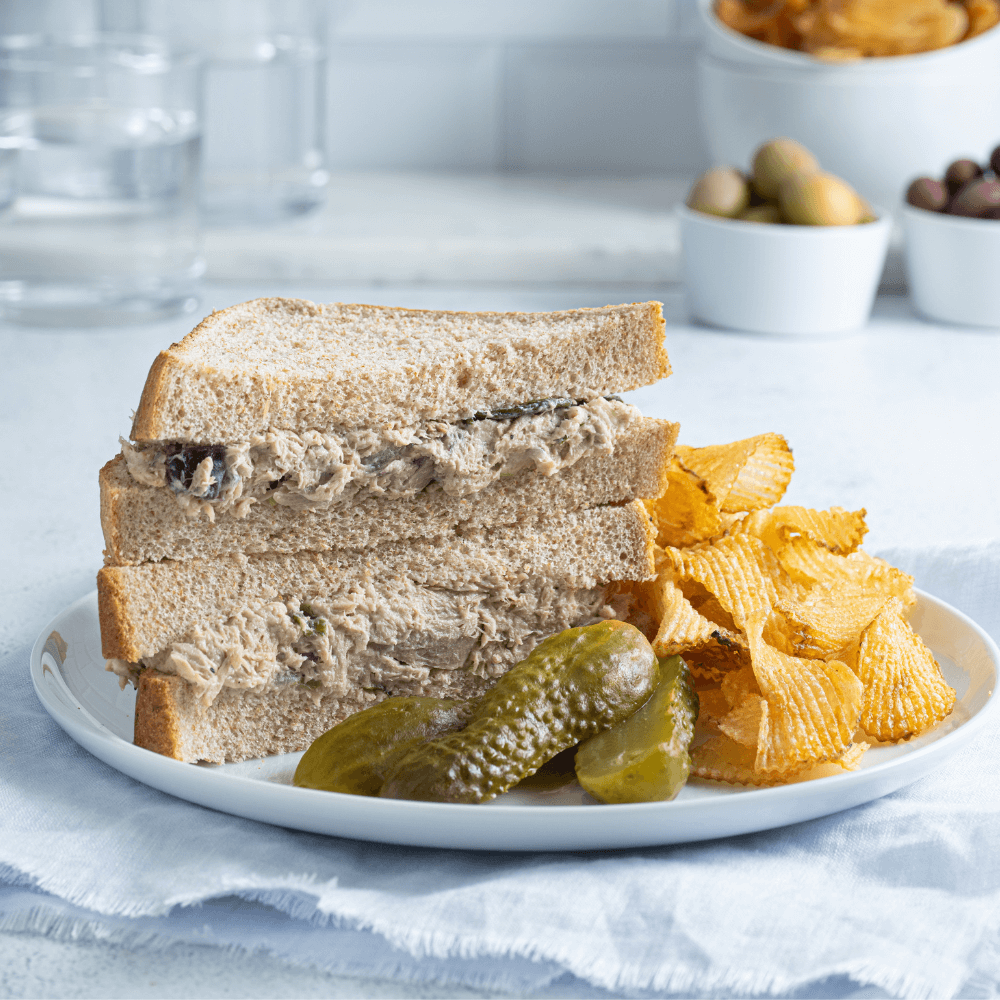 White plate with two halves of a tuna salad sandwich stacked on top of one another. On the plate are three pickle slices and a handful of crinkle-cut potato chips.