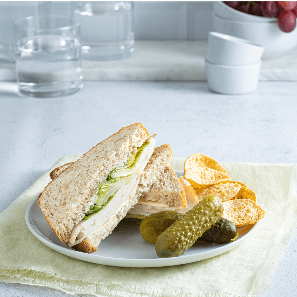 White plate with a sliced turkey & Swiss cheese sandwich on it, beside two pickles and handful tortilla chips.