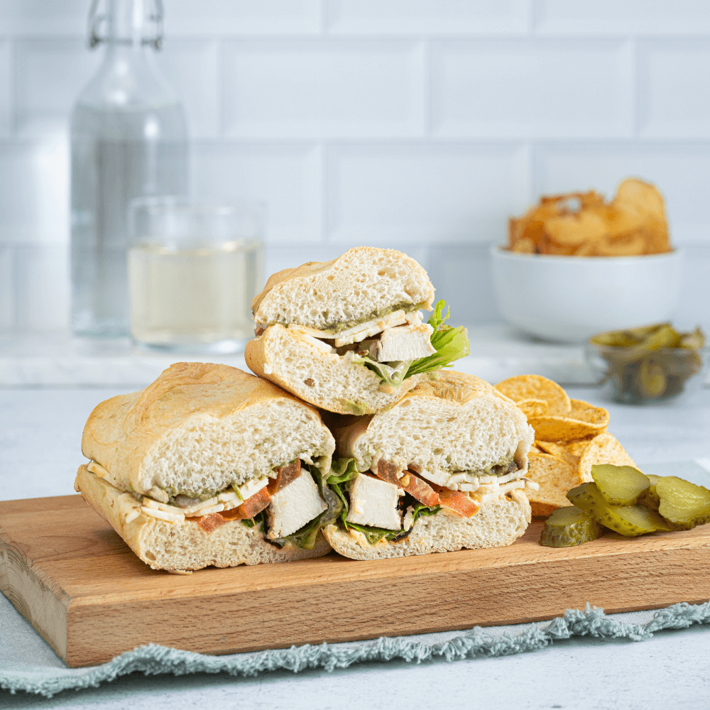 On a wooden cutting board are three segments of a chicken sub. Beside it are sliced pickles and tortilla chips.