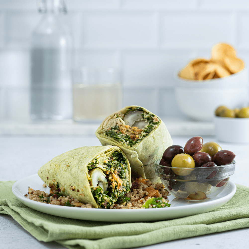 A white plate is on a pale green napkin. On the plate is a falafel wrap cut in half. Beside it is a small clear glass bowl of mixed olives.