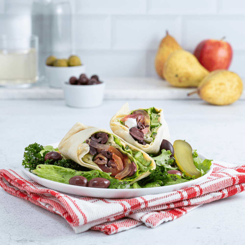 A white plate is on a red and white plaid cloth. On the plate is a Greek salad wrap cut in half on a bed of mixed greens with olives and sliced pickles.