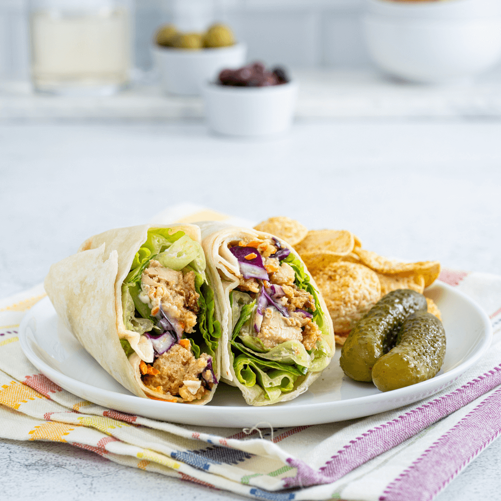 A white plate is on a rainbow-striped cloth. On the plat is a teriyaki chicken wrap cut in half, Red cabbage, lettuce, carrots, and chicken are visible. Beside it are two pickles and a handful of tortilla chips.