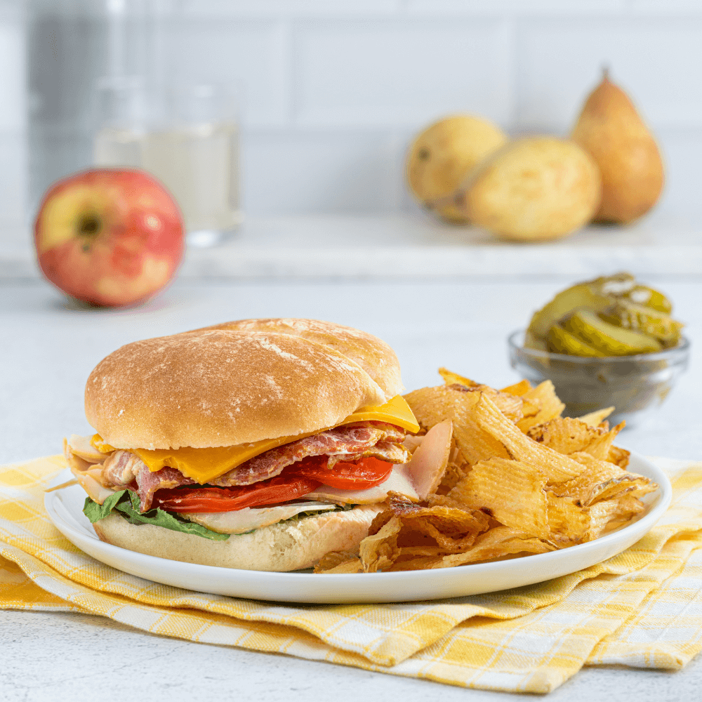 A white plate on a yellow checked dishcloth. On the plate is a turkey club sandwich with cheese, ham, turkey, lettuce, and tomatoes. Beside it are crinkle-cut potato chips.