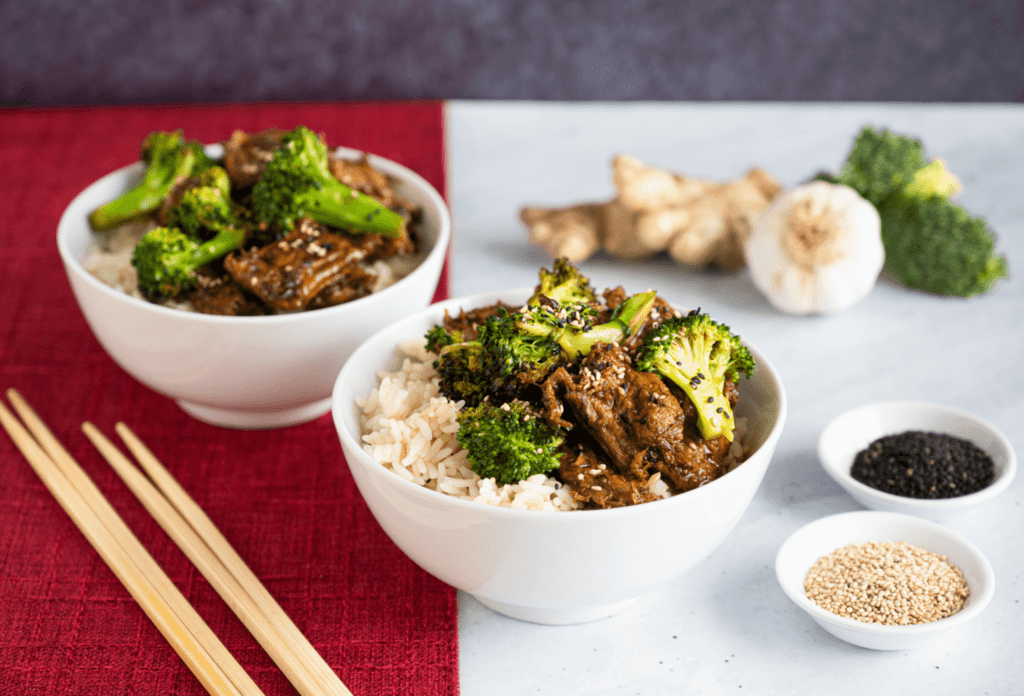 Two white bowls of rice with plant-based beef and broccoli next to two pairs of chopsticks.