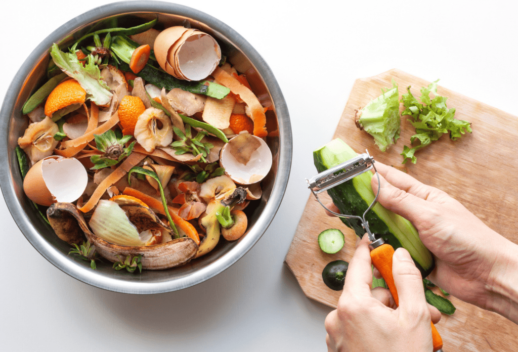 Female hands brush cucumber with knife near iron bowl with organic food waste. Vegetable peelings and food leftovers ready to compost. Ecological, recycling waste concept.