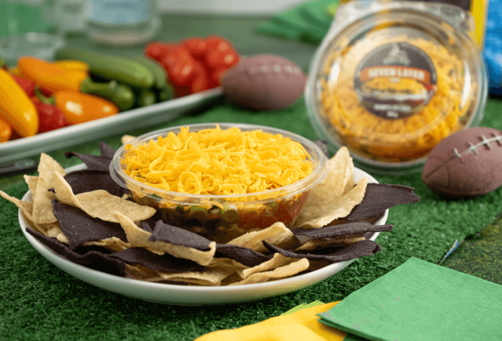 PLate with blue and white corn tortilla chips and a bowl of seven layer dip in the centre. In the background are assorted fresh vegetables and crudites.