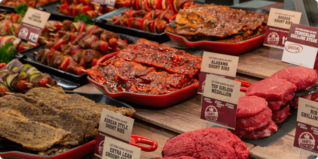 A close-up of a refrigerated display case in a Farm Boy store meat department. Various cuts of beef are packaged on trays, with labels indicating the cut and origin. Signs advertise "Canadian beef" and "100% extra lean ground beef.