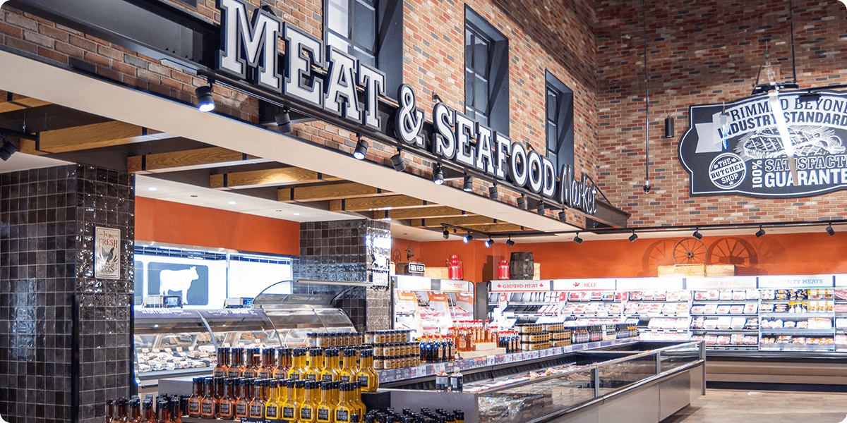 A well-lit Farm Boy store meat and seafood department with a variety of fresh meats, fish, and shellfish on display in refrigerated cases. Signs above the cases advertise that the department meets industry standards and offers a satisfaction guarantee. There is also a butcher counter visible in the image.