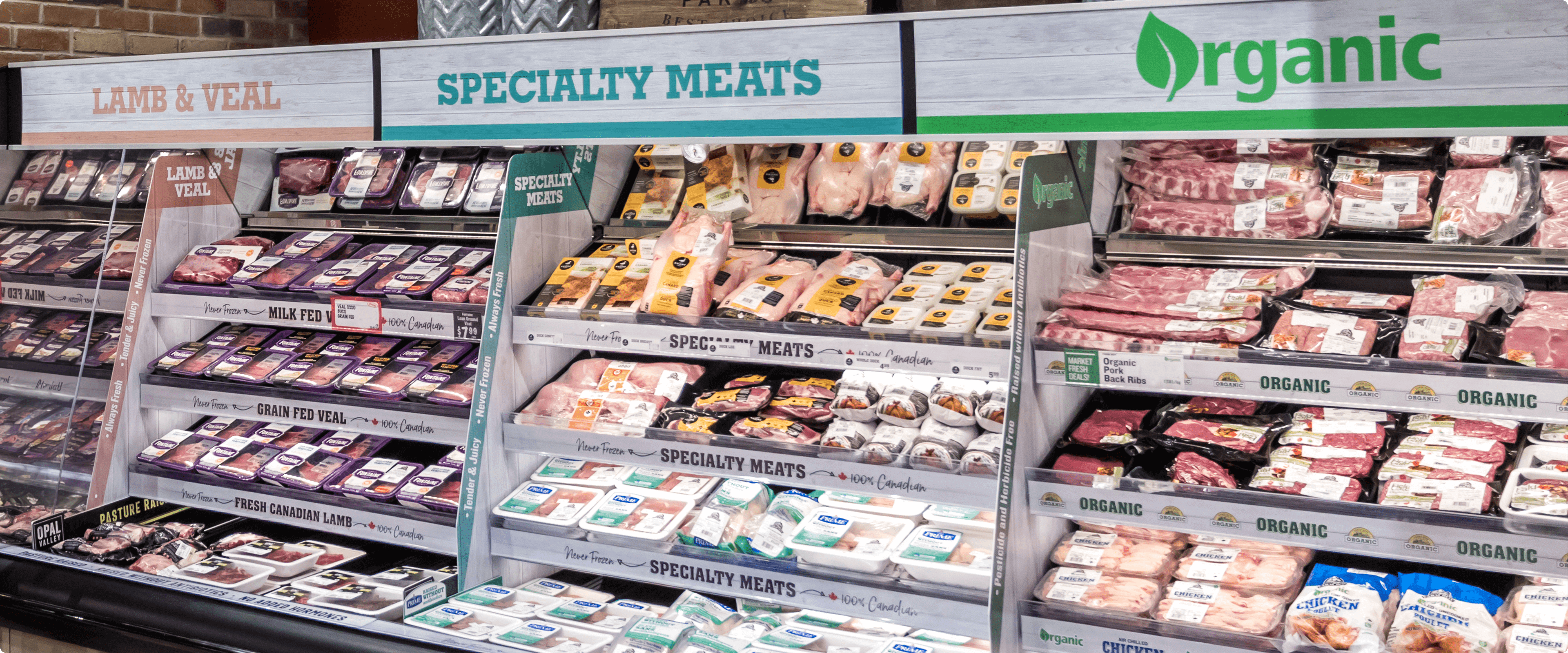 A well-lit section of a Farm Boy store meat department with a variety of fresh meats on display in refrigerated cases. Signs above the cases advertise organic and specialty meats, including lamb, veal, and grain-fed veal.