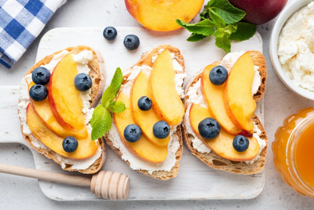 A square plate containing a slice of toast with ricotta cheese, honey, peaches, and blueberries. The toast is a thick slice of golden brown bread. A generous amount of ricotta cheese is spread across the toast. Honey is drizzled across the ricotta cheese in a decorative pattern. Sliced peaches and blueberries are scattered on top of the toast. The peaches are light orange in colour with a slight reddish hue near the skin. The blueberries are a dark blue colour.
