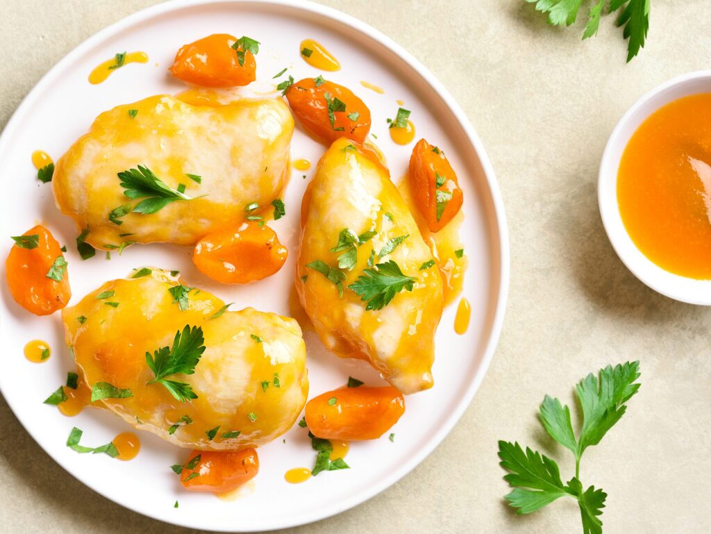 A pan-seared chicken breast with a peach sauce glaze on a white plate. There are also  carrot sticks on the plate. 