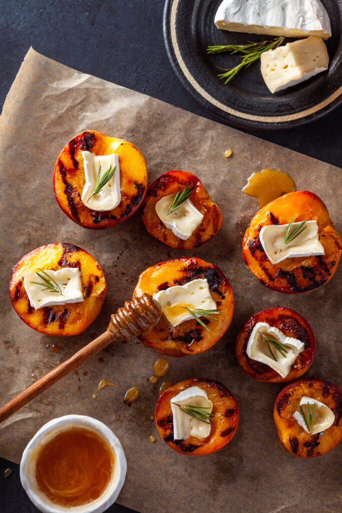 A plate of grilled peaches with brie, rosemary, and honey. The halved peaches are golden brown with grill marks. Slices of brie cheese rest on top of the peach halves. A sprig of fresh rosemary is positioned diagonally across the plate. There is a drizzle of honey across the peaches and brie.
