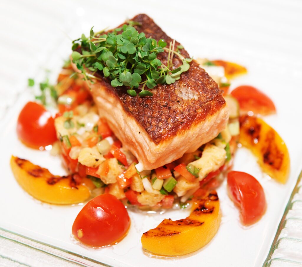 A white plate with a pan-seared salmon fillet in the center. The salmon has a crispy, golden-brown crust. To the right of the salmon are slices of grilled summer vegetables, including zucchini, yellow squash, and red bell pepper. There are also two grilled peach halves to the left of the salmon. All the vegetables and peaches have grill marks and a slightly charred appearance. A sprig of fresh rosemary is placed diagonally across the salmon fillet.
