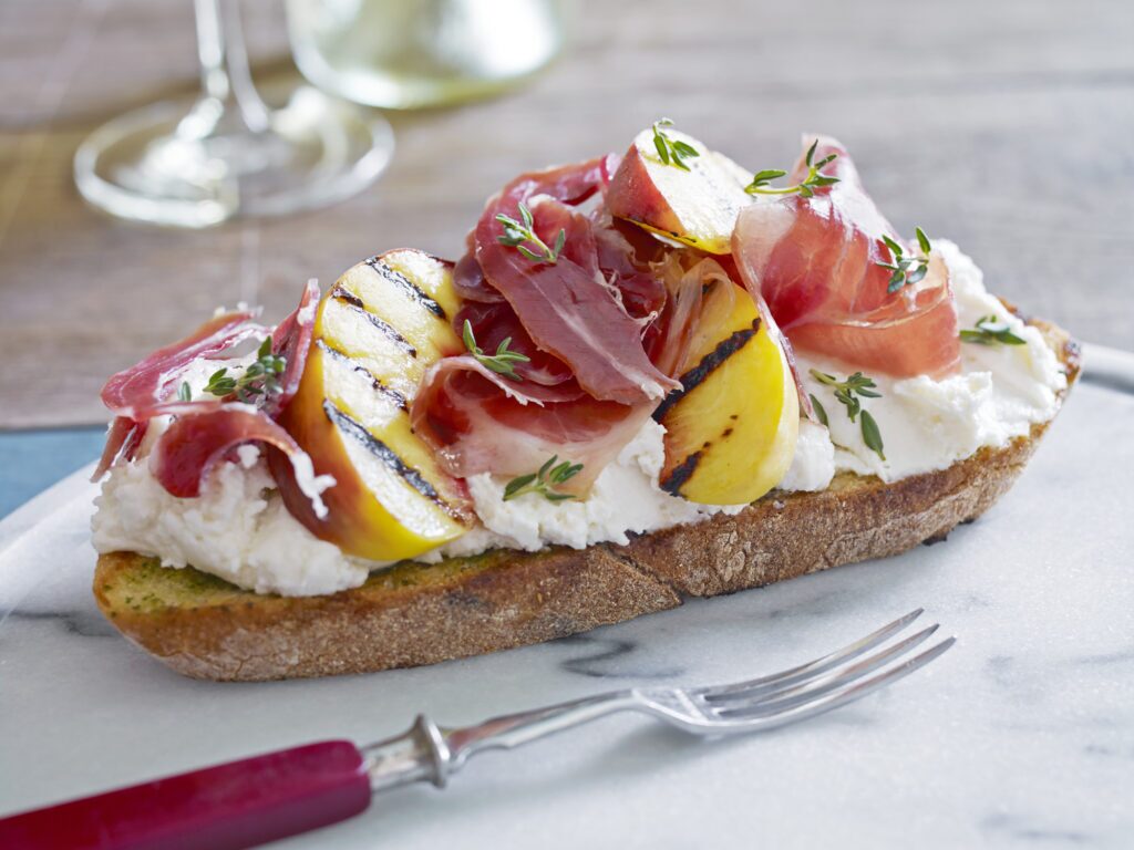 A close-up image of a crostini appetizer on a wooden cutting board. The crostini is a small slice of toasted bread topped with thinly sliced prosciutto and sliced peaches. The prosciutto is a cured ham that is pink and fatty. The peach slices are light orange in color and some appear riper with a slight reddish hue. A single green colored fork with a metal tine rests beside the crostini on the cutting board. In the background, slightly blurred, is the edge of a light coloured counter top.