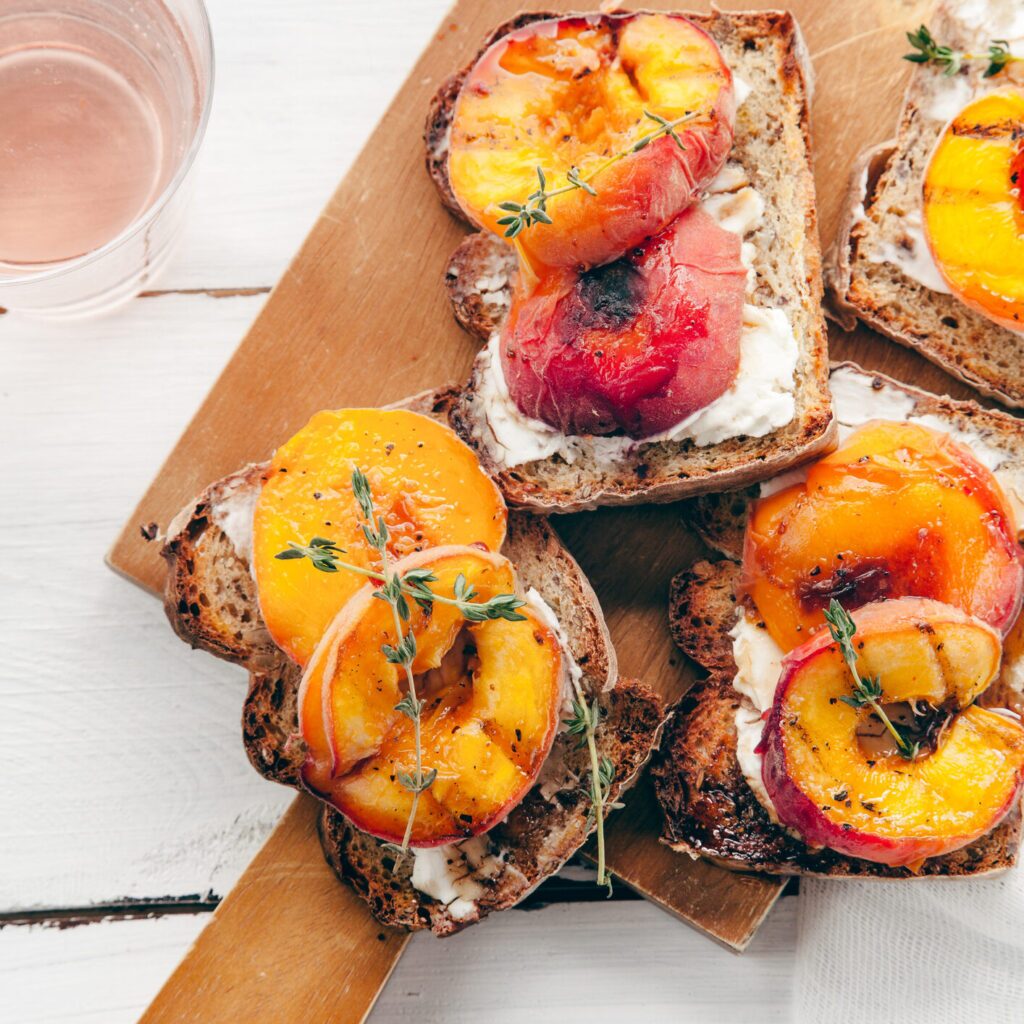 A plate of toast with cream cheese, thyme, and peaches. The toast is two slices of golden brown bread. There is a thick spread of cream cheese on both slices of toast. Several sprigs of fresh thyme are scattered on the cream cheese. Two peach halves are placed on top of the toast. The peach halves are sliced thinly and are light orange in colour with a slight reddish hue near the skin.